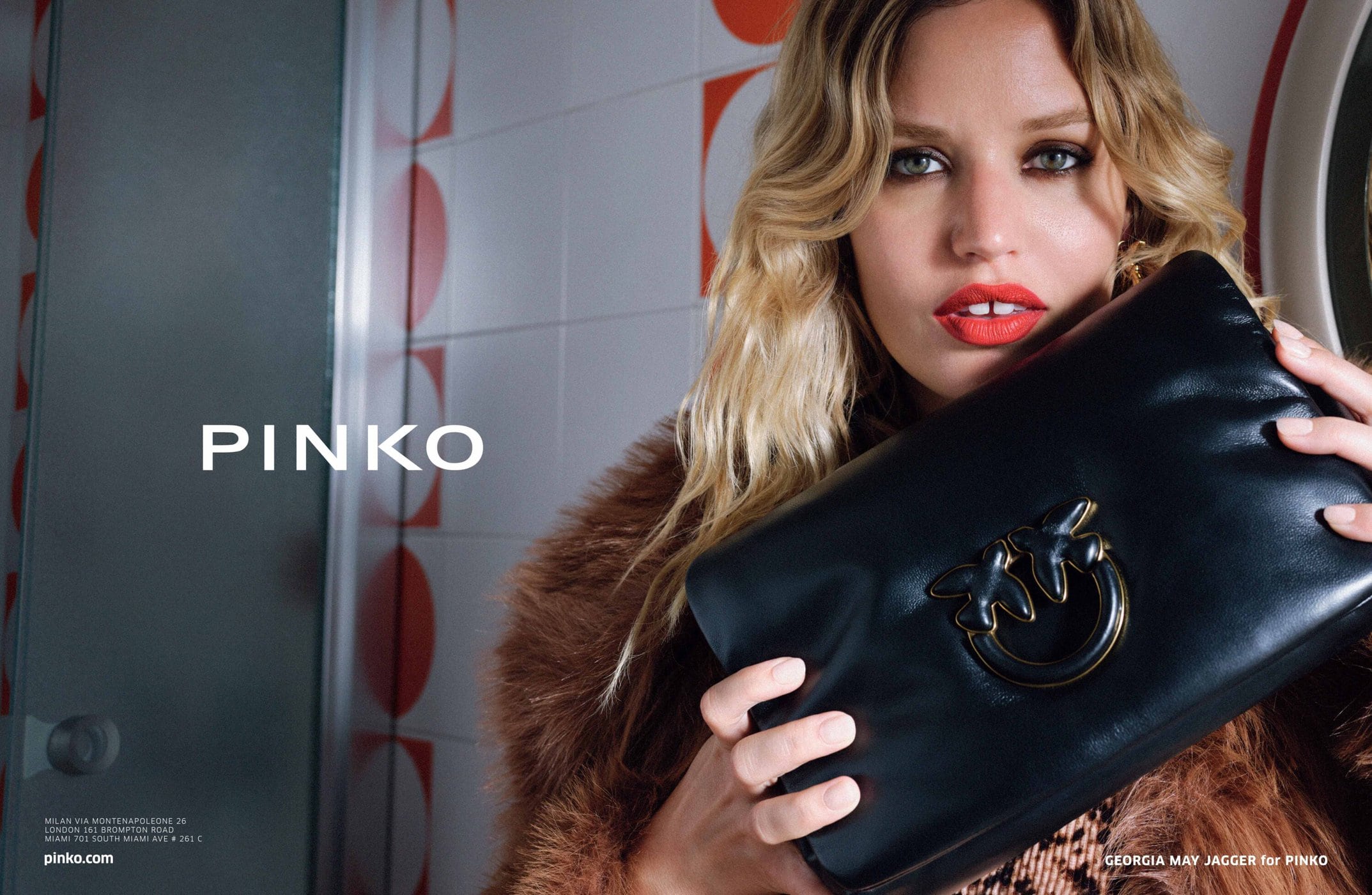 Blonde woman presents a black Pinko clutch with metal detail in a fashion ad, wearing a fur coat and red lipstick against a patterned wall.