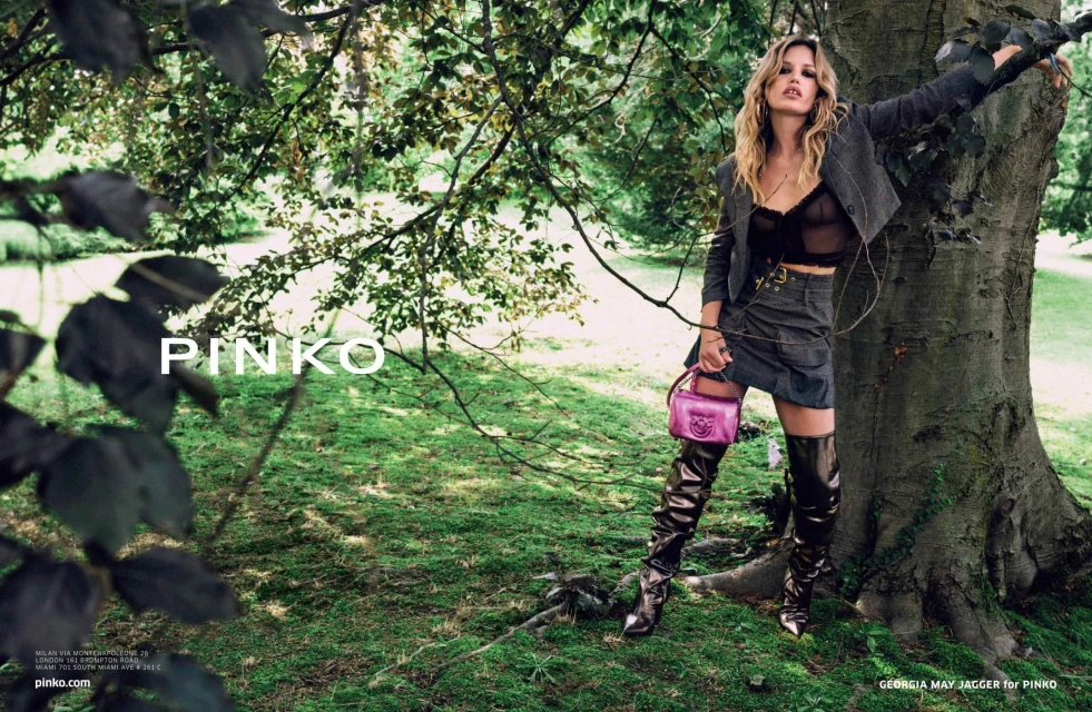 Fashion shoot in wooded area featuring a blond adult woman wearing a dark jacket, miniskirt, high boots, and pink purse. 