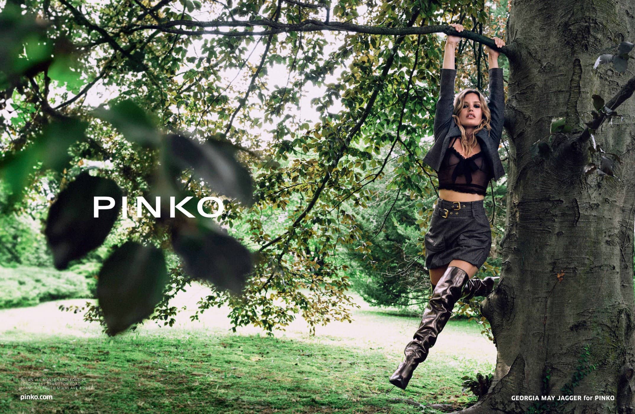 A fair-skinned adult woman in black top, shorts, jacket, and tall shiny boots hangs from a tree branch in a park-like setting.