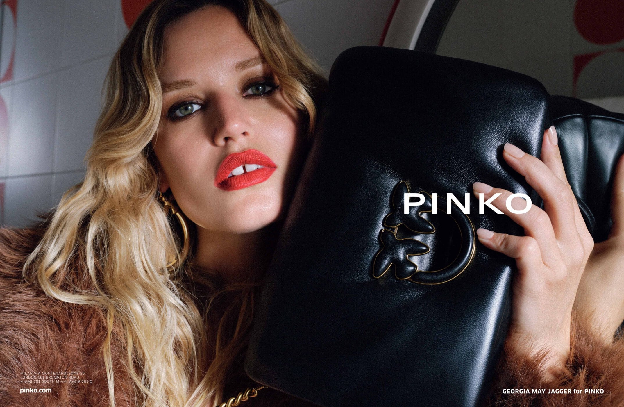 Close-up of a blonde adult woman posing with a black leather Pinko handbag in a studio, wearing a fur coat and bright red lipstick.