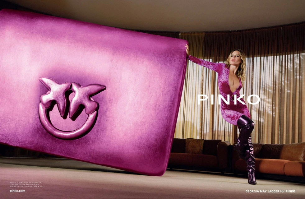Pinko fashion campaign featuring Georgia May Jagger in a pink catsuit, leaning against an oversized Pinko handbag in a studio setting.