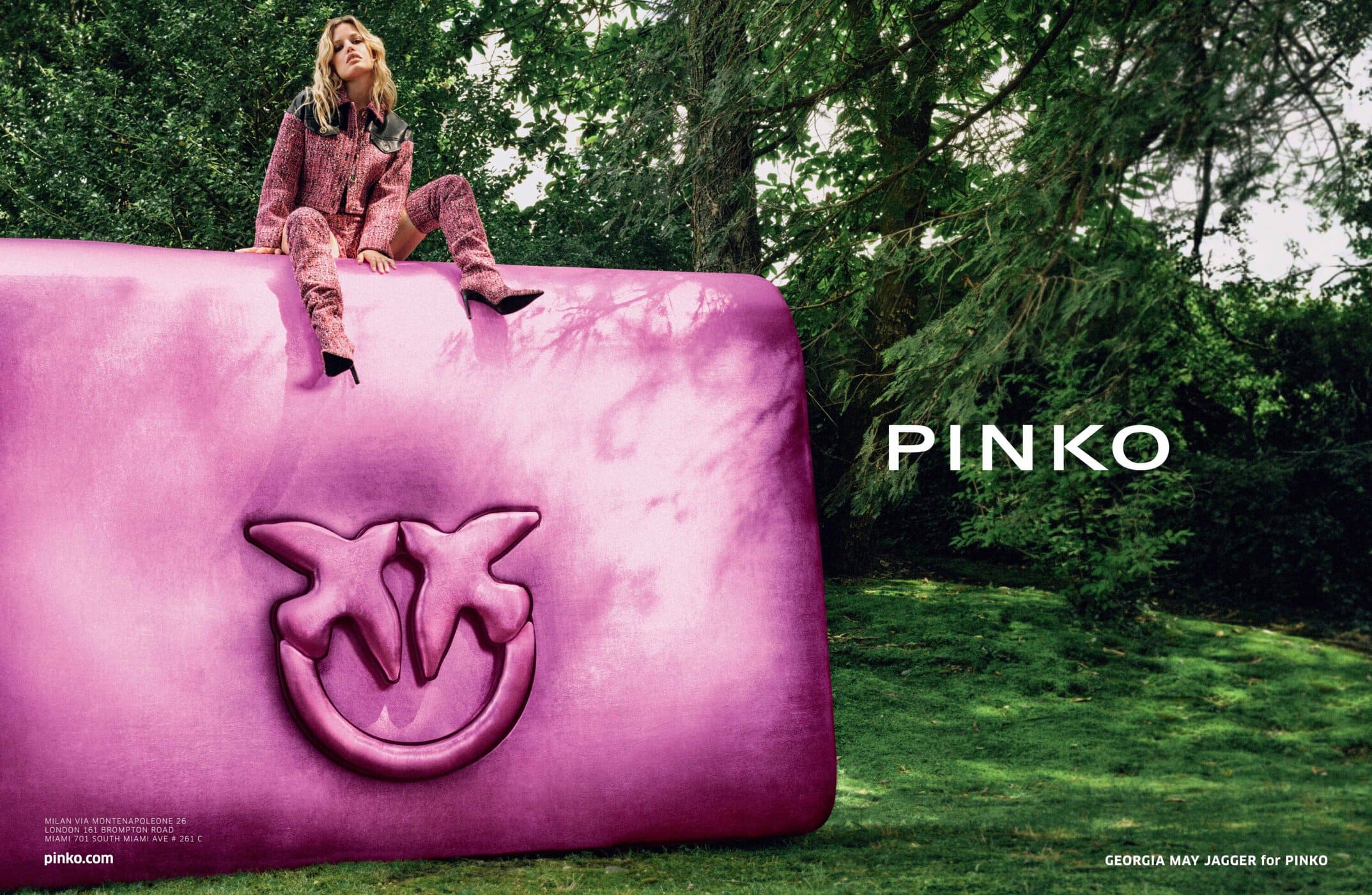 Fashion shot featuring an adult female model sitting on a giant pink Pinko bag in a grassy outdoor setting with trees in the background.