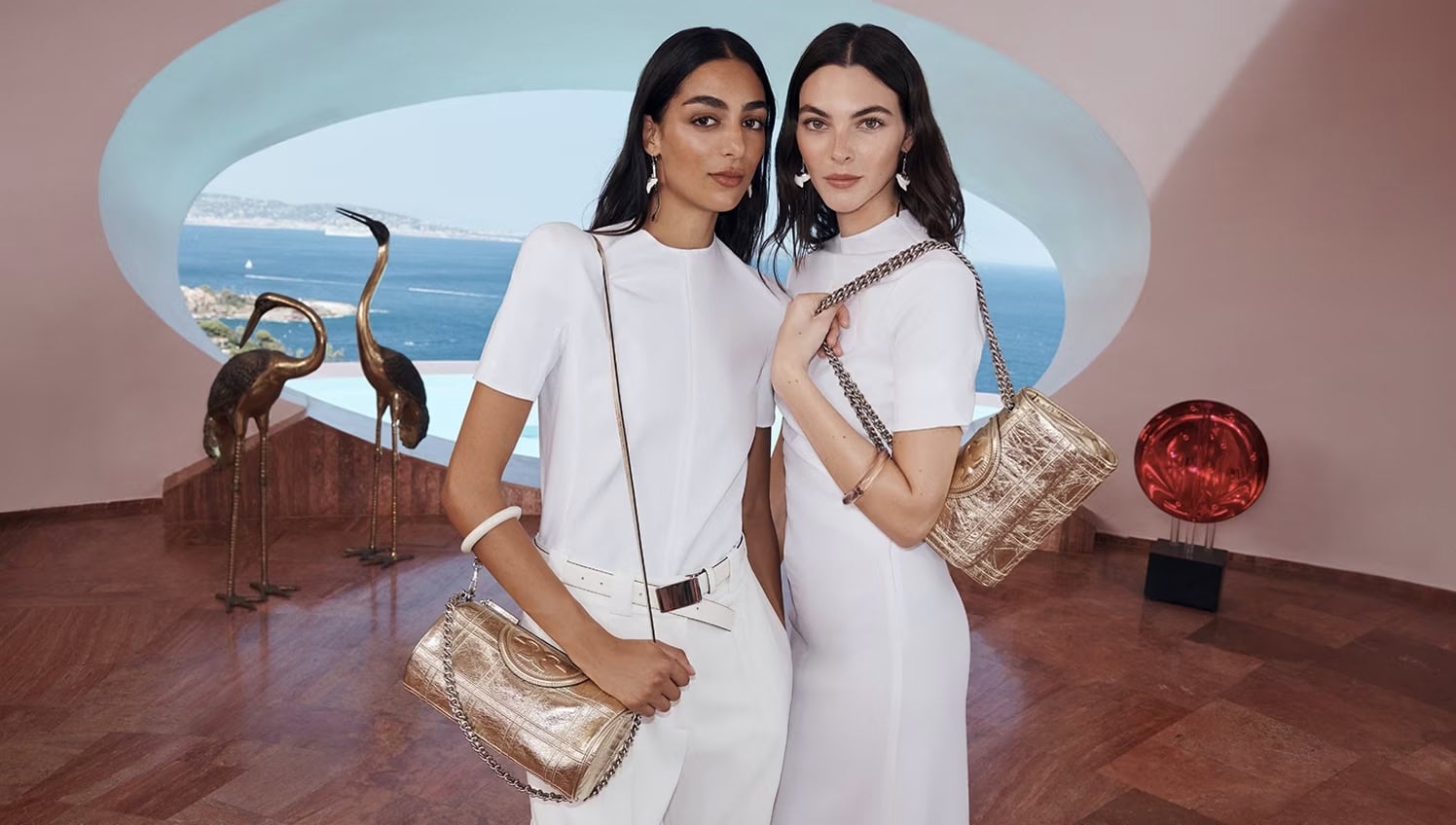 Two women in white outfits pose indoors with gold purses, sculptural decor, and a coastal view through a rounded window.