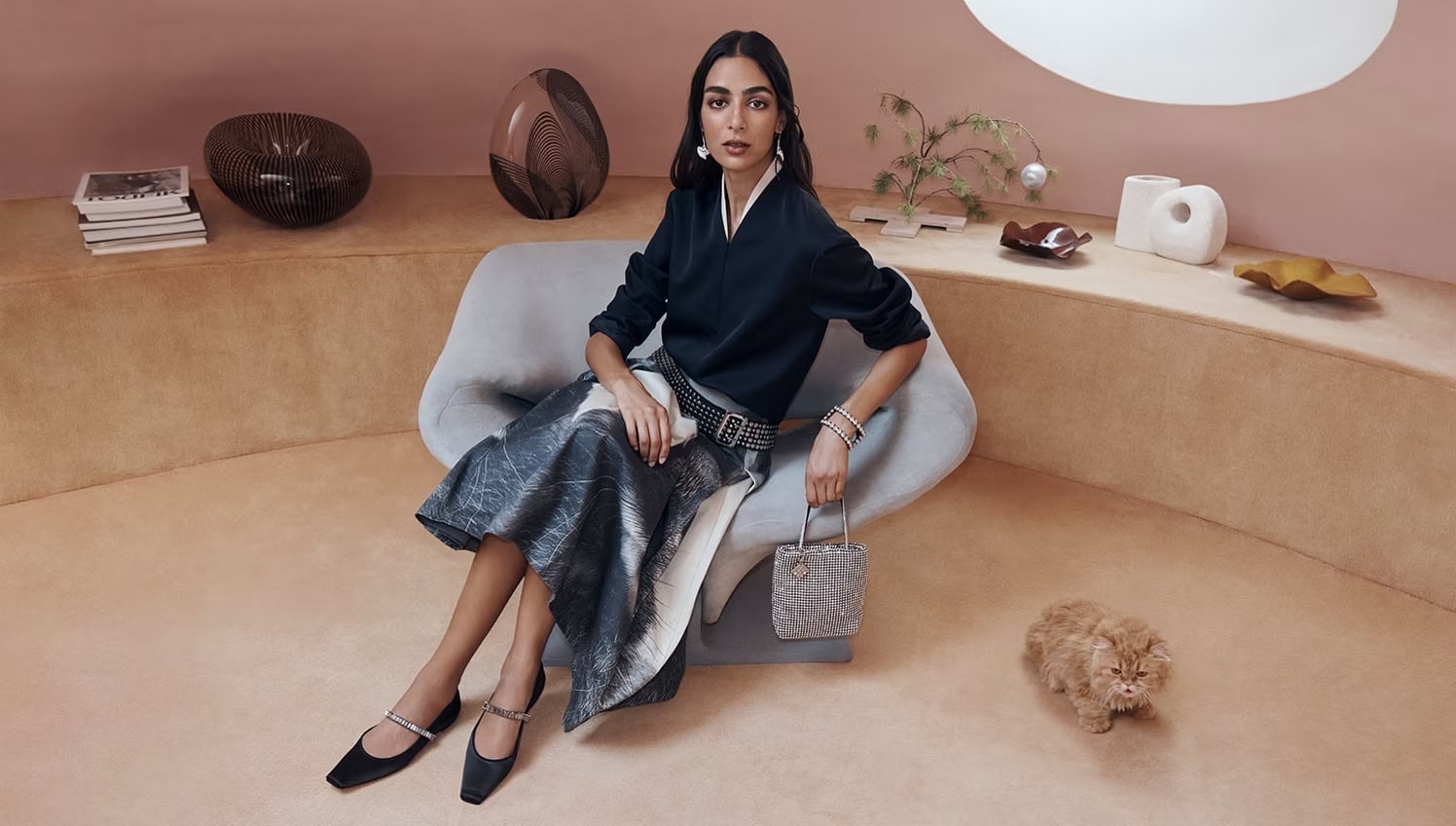 Elegant woman in designer dress and accessories poses indoors with an orange kitten, showcasing fashion and interior design elements.