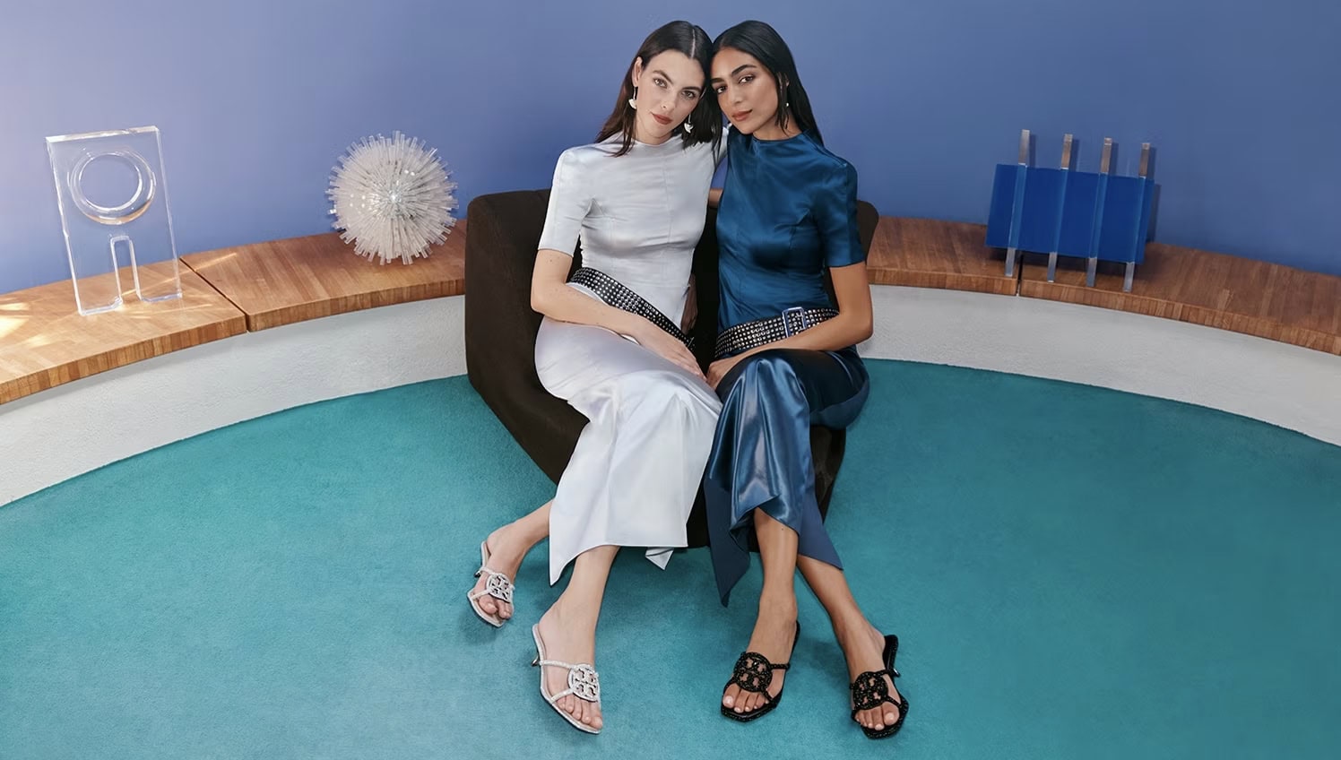 Two women in satin outfits pose seated in a studio with a turquoise floor, wearing elegant heels and belts, under bright lighting.