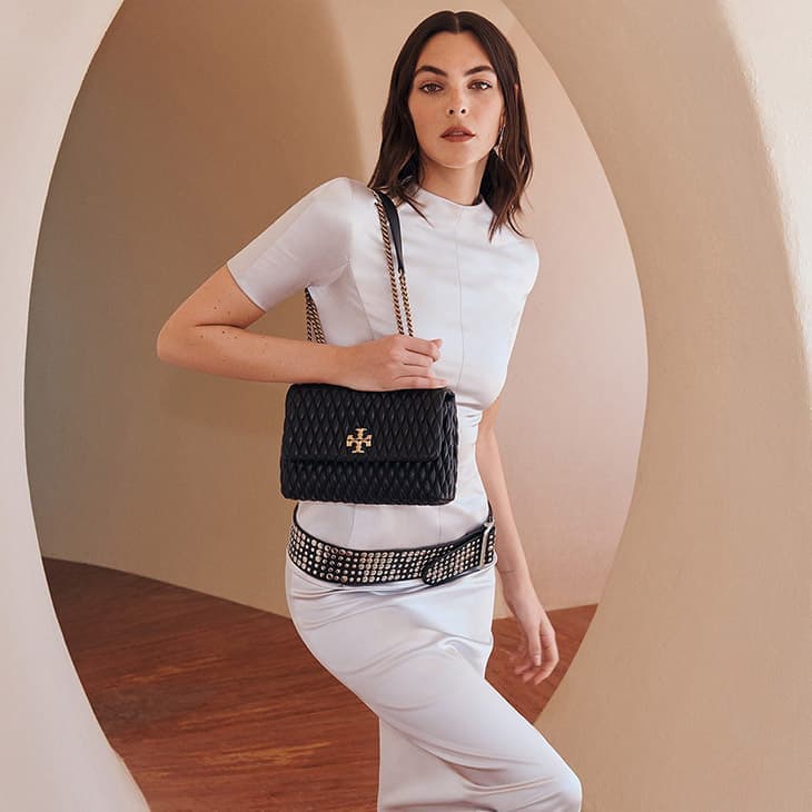 Elegant woman in a sheath dress poses indoors, showcasing a quilted leather handbag with chain strap and studded belt, fashion editorial.
