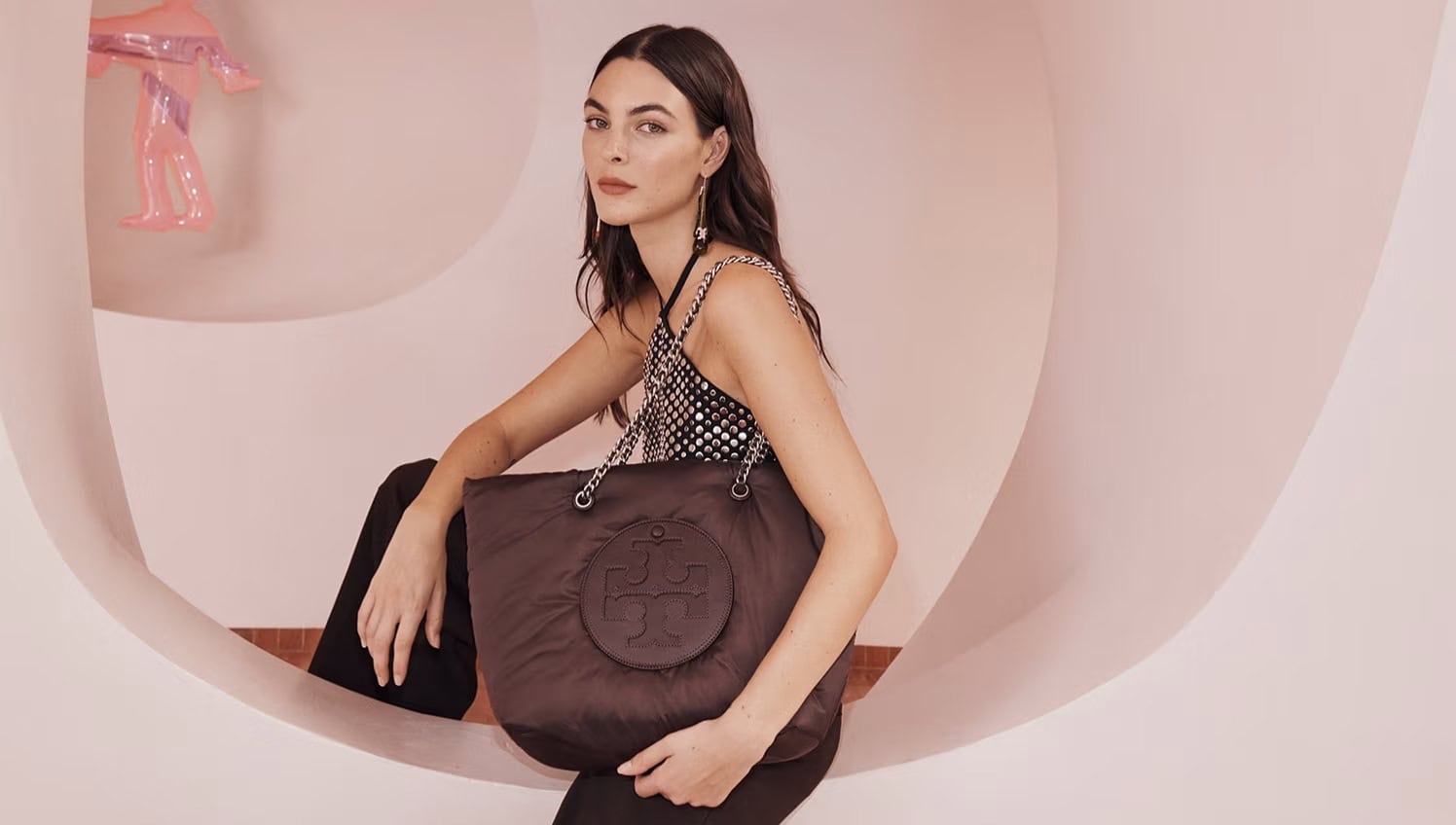 Brunette woman poses indoors with a brown Tory Burch tote bag with chain straps, wearing a sparkly top and dark pants.