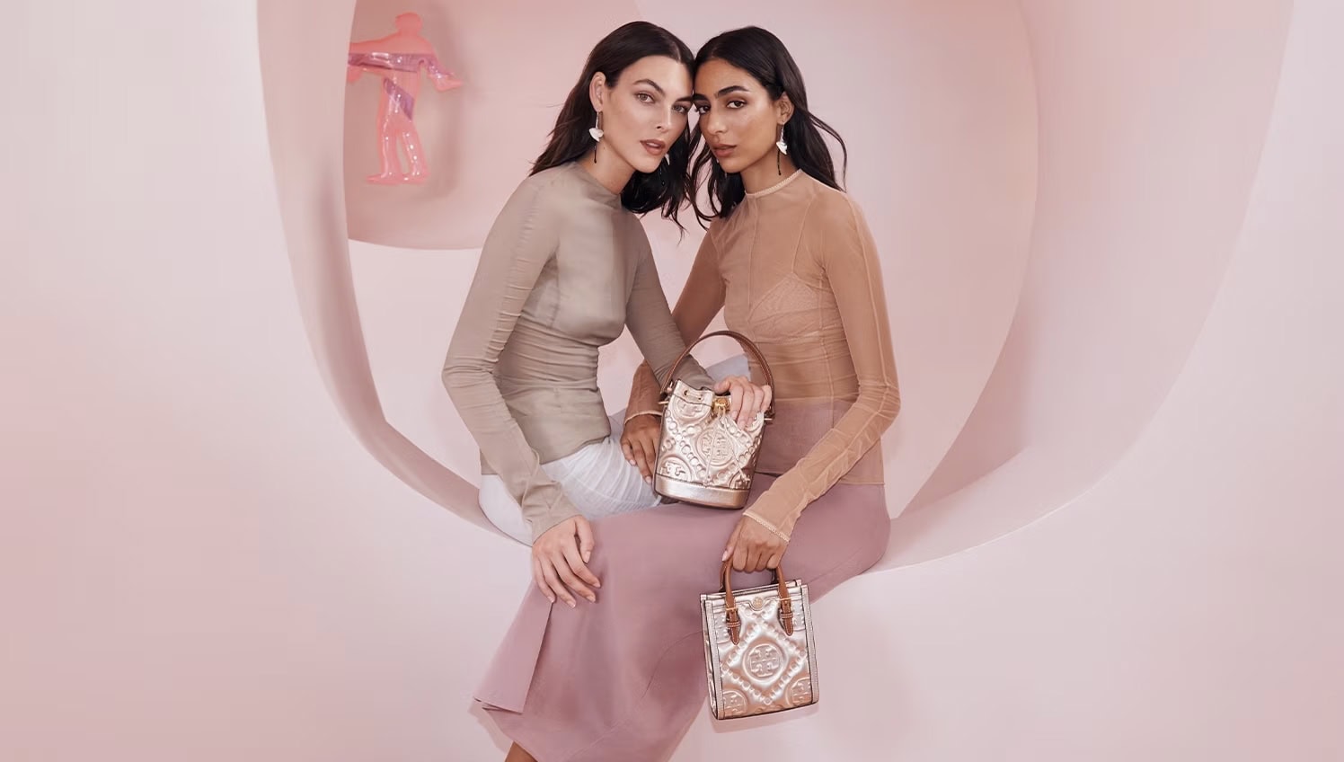 Two women pose indoors in sheer tops, holding ornate handbags against a soft pink backdrop, creating an elegant, fashionable composition.