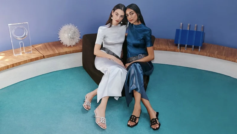 Two female models seated together on a dark armchair wearing matching satin outfits and studded belts against a blue wall.