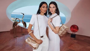 Two female models in white outfits holding gold handbags inside a room overlooking the sea for Tory Burch Holiday 2023.