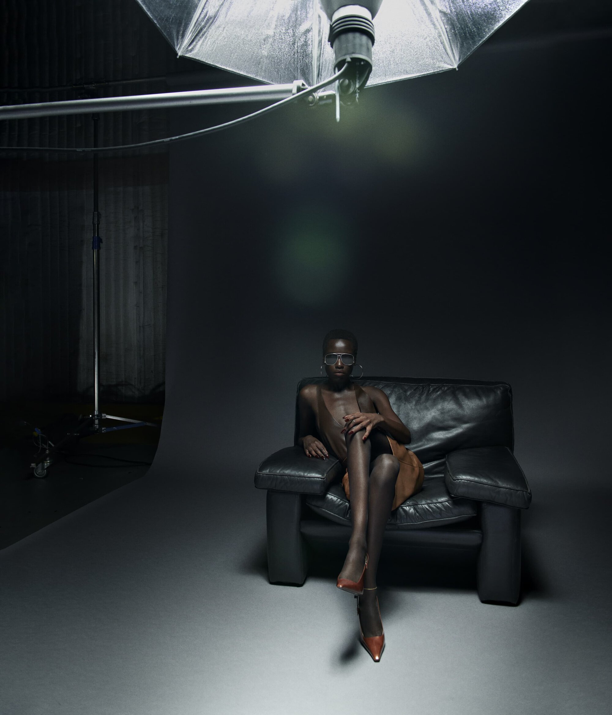 A slender dark-skinned adult woman sits on a black leather armchair in a photo studio with silver umbrella lighting and neutral backdrop.