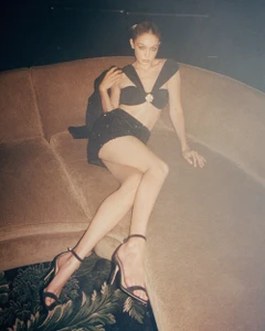 Gigi Hadid seated on a curved brown sofa wearing a black two-piece set and heels for Self-Portrait.