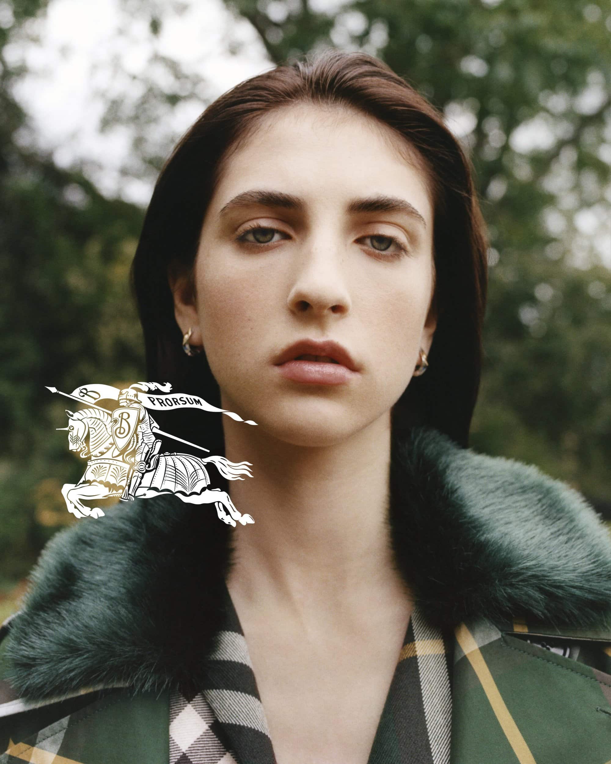 Close-up of a woman with brown hair and a plaid coat in an outdoor setting, overlaid with the Burberry logo. Soft, diffused lighting.