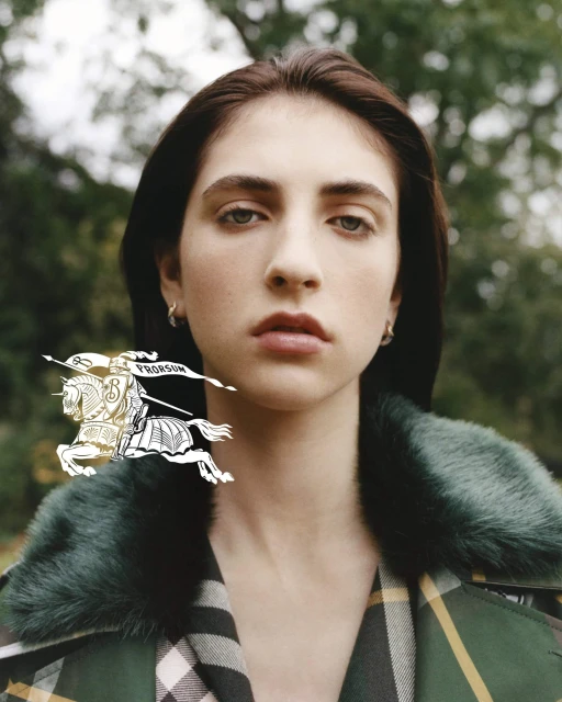 Close-up of a woman with brown hair and a plaid coat in an outdoor setting, overlaid with the Burberry logo. Soft, diffused lighting.