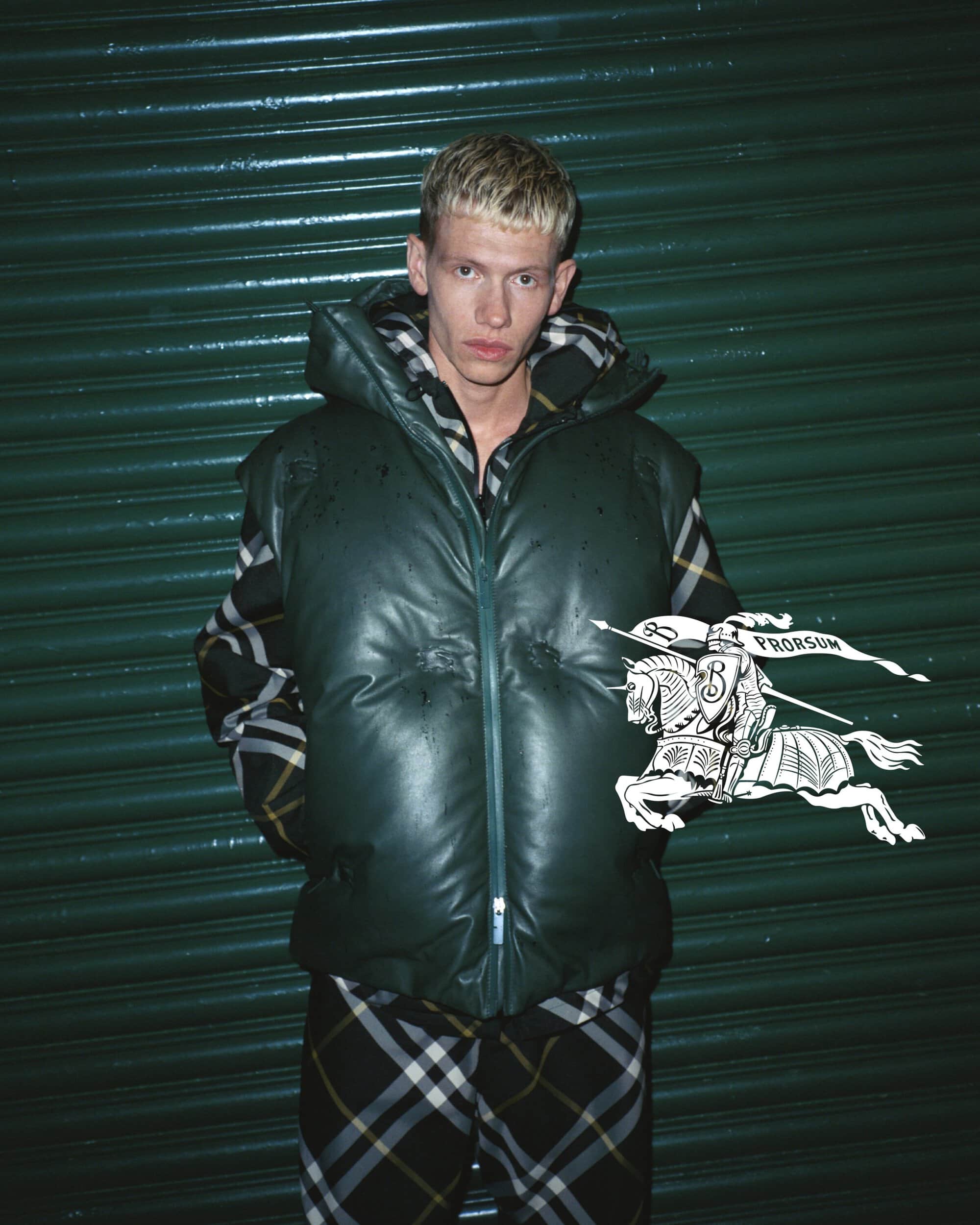 A blond adult male poses in a green, hooded puffer vest and plaid pants, against a green roll-up door, showcasing a modern streetwear style.