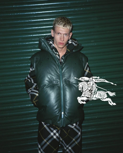 A blond adult male poses in a green, hooded puffer vest and plaid pants, against a green roll-up door, showcasing a modern streetwear style.