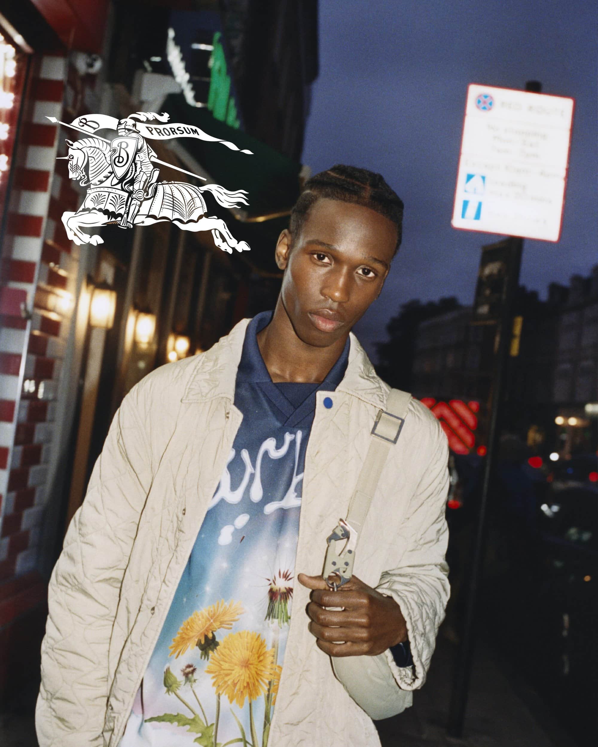 Young adult posing outdoors at dusk. He is wearing a graphic tee and a light beige jacket with a crossbody bag. Pedestrian crossing sign.