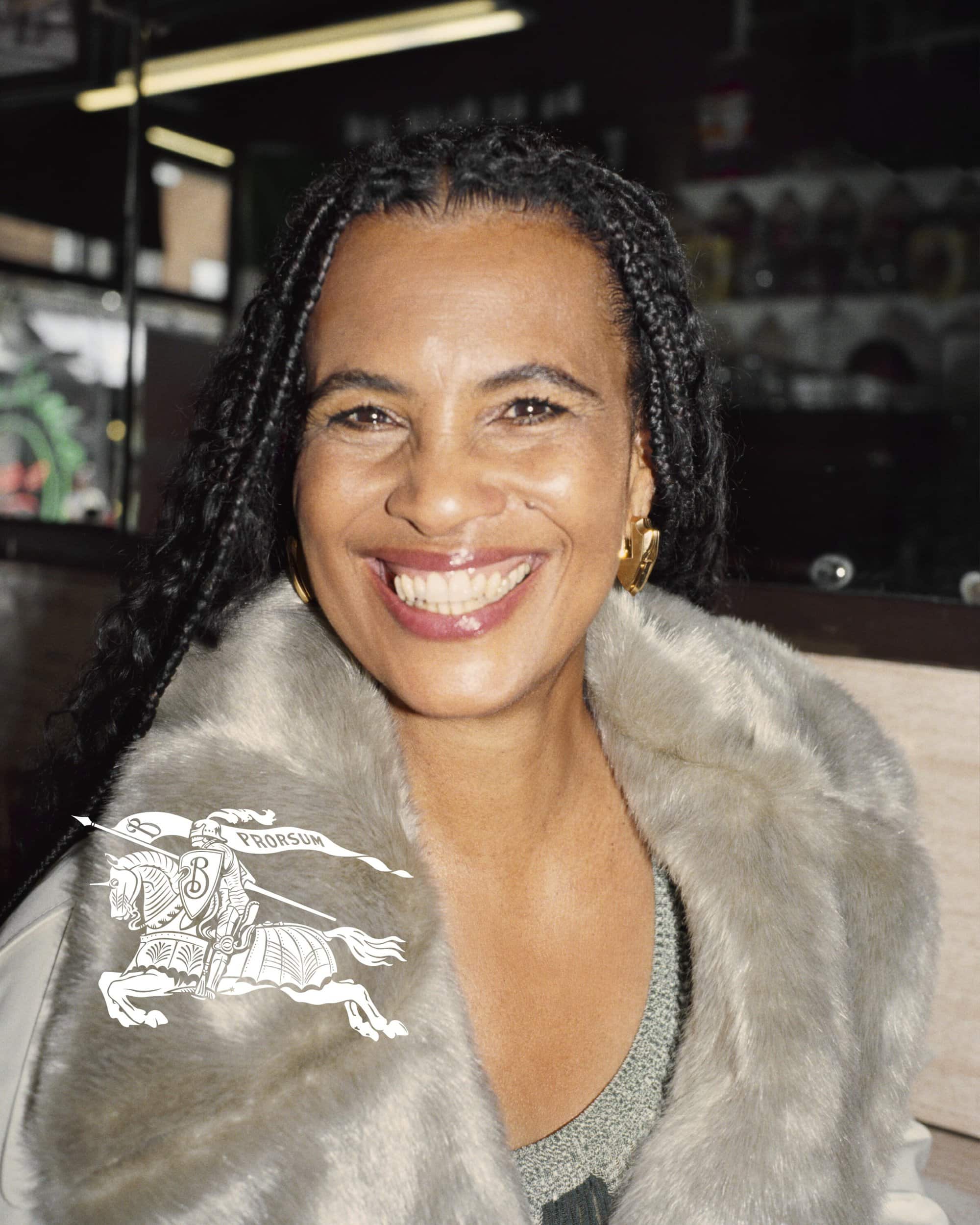 Smiling adult woman in a gray faux fur coat and earrings. She has long black braided hair and is indoors with a dark background.