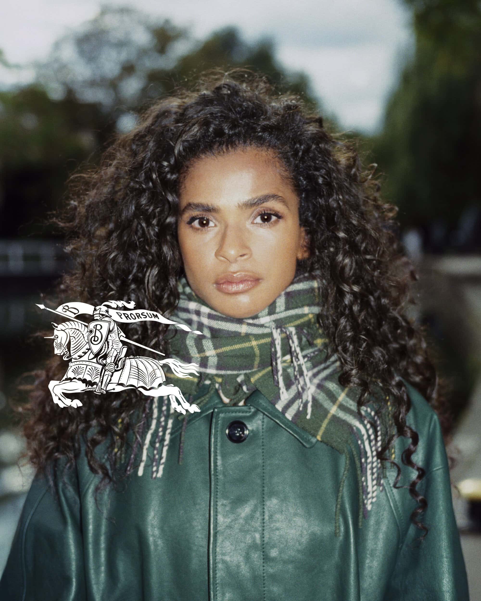 Brunette woman with curly hair poses wearing a green coat and plaid scarf outdoors with a river and trees in the background.