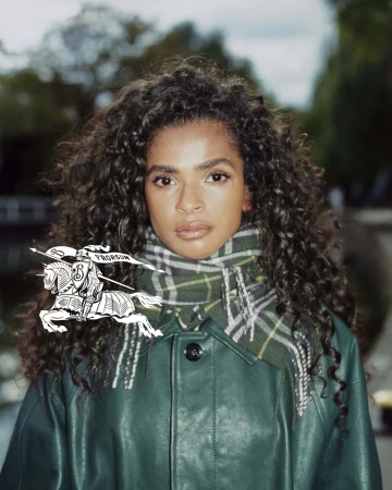 Brunette woman with curly hair poses wearing a green coat and plaid scarf outdoors with a river and trees in the background.