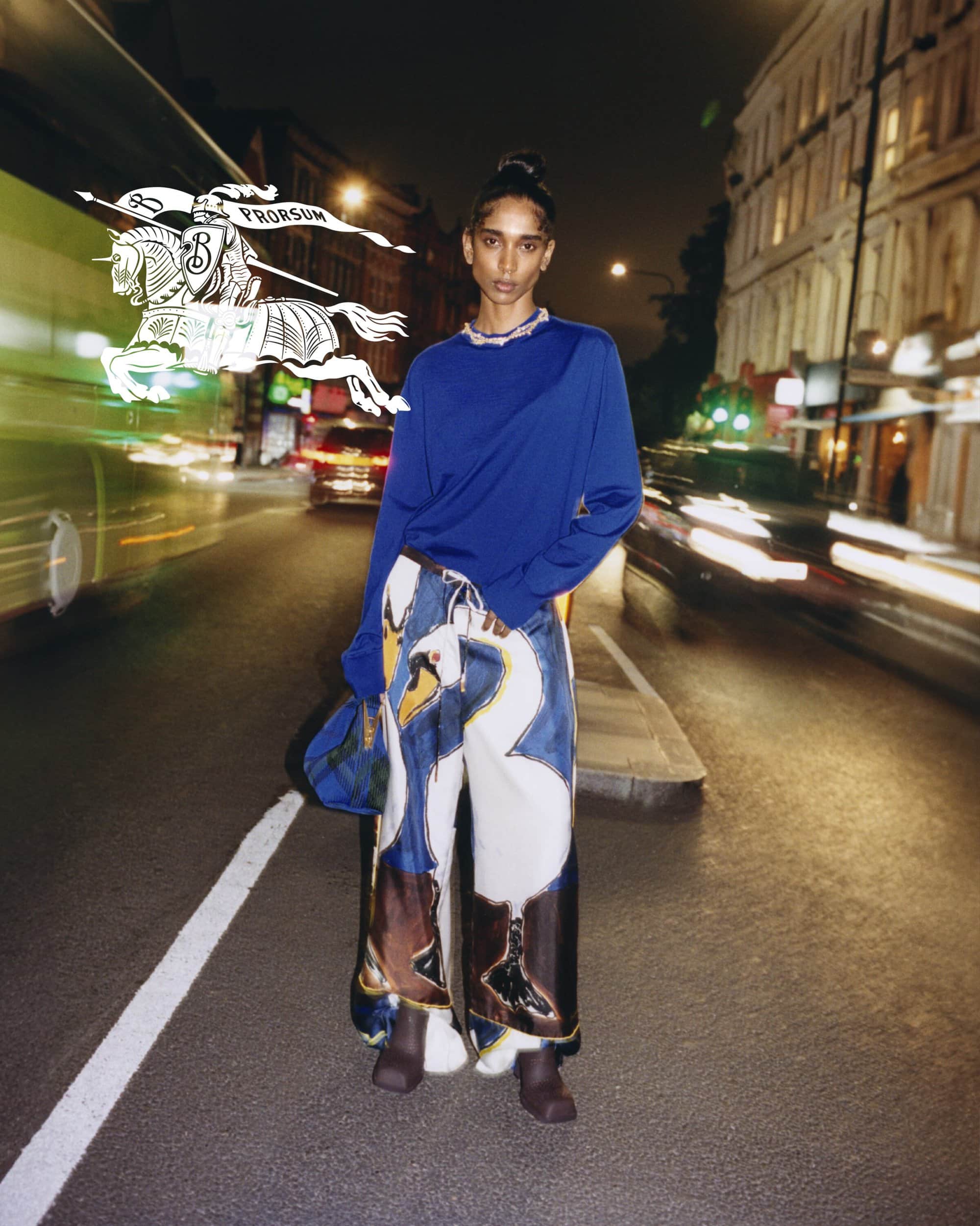 Woman poses on city street at night in an blue top and pants with swan designs, with bus and blurred vehicle lights in the background.