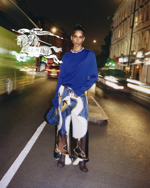 Woman poses on city street at night in an blue top and pants with swan designs, with bus and blurred vehicle lights in the background.