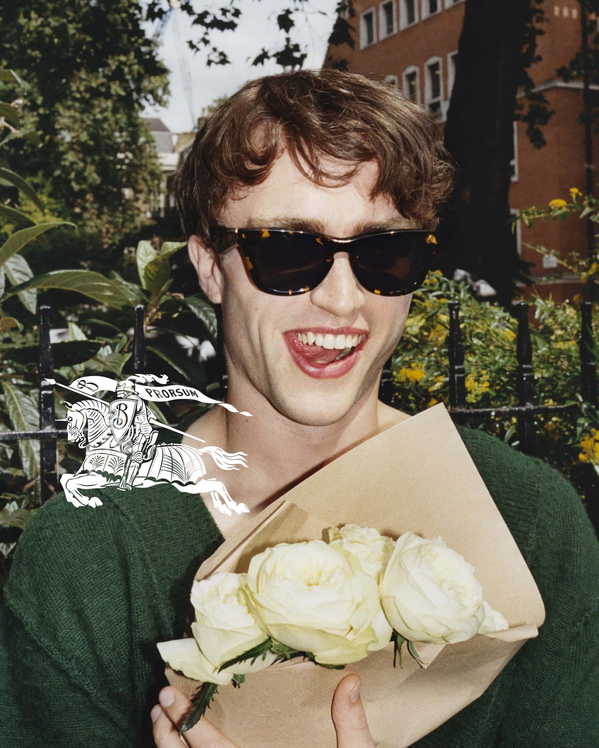 Young man in sunglasses holds a bouquet of white roses. Outdoors in a park with buildings in the background. Fashion editorial style.