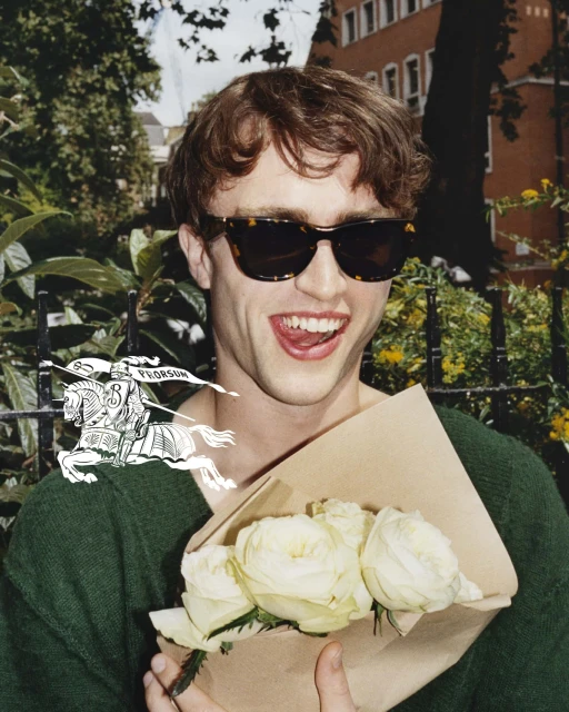 Young man in sunglasses holds a bouquet of white roses. Outdoors in a park with buildings in the background. Fashion editorial style.
