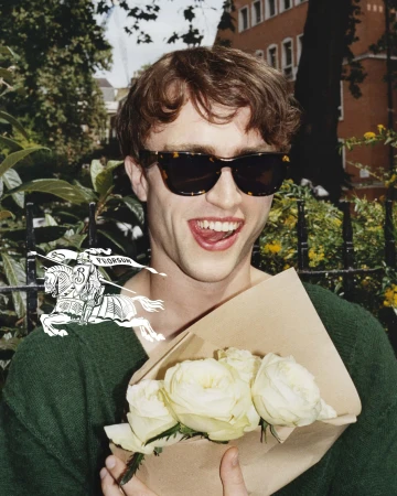 Young man in sunglasses holds a bouquet of white roses. Outdoors in a park with buildings in the background. Fashion editorial style.