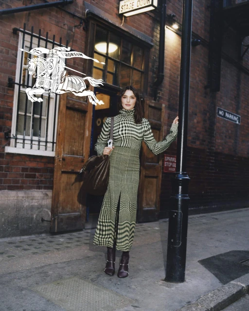 An adult female stands on a city street in a patterned dress and brown boots, holding a large bag, brick building behind her.