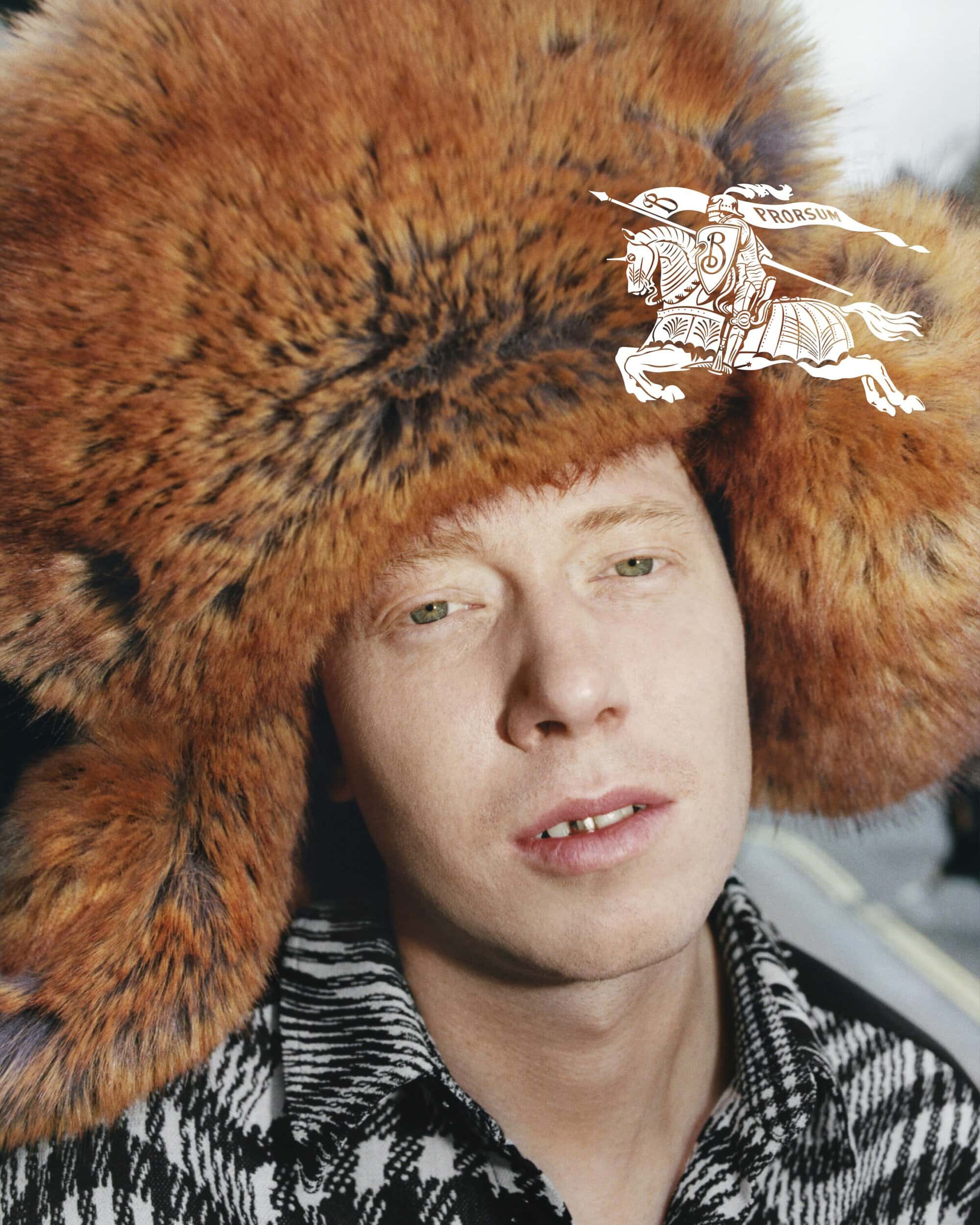 Portrait of a man with fair skin and ginger hair, wearing a fur hat and a patterned shirt, looking at the camera.