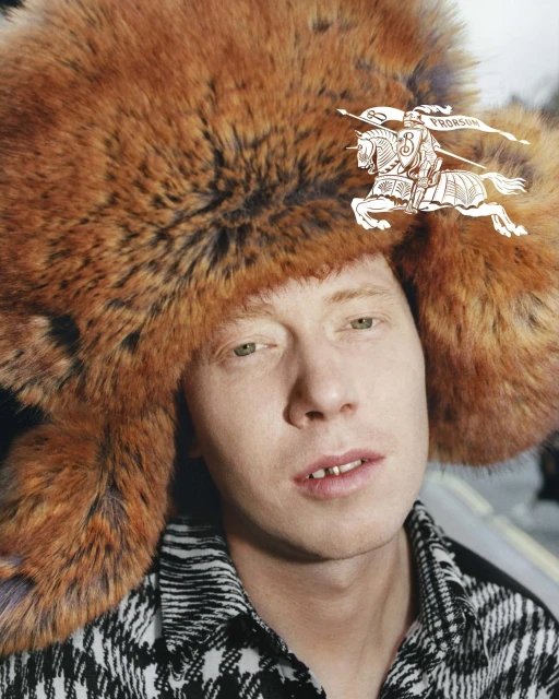 Portrait of a man with fair skin and ginger hair, wearing a fur hat and a patterned shirt, looking at the camera.