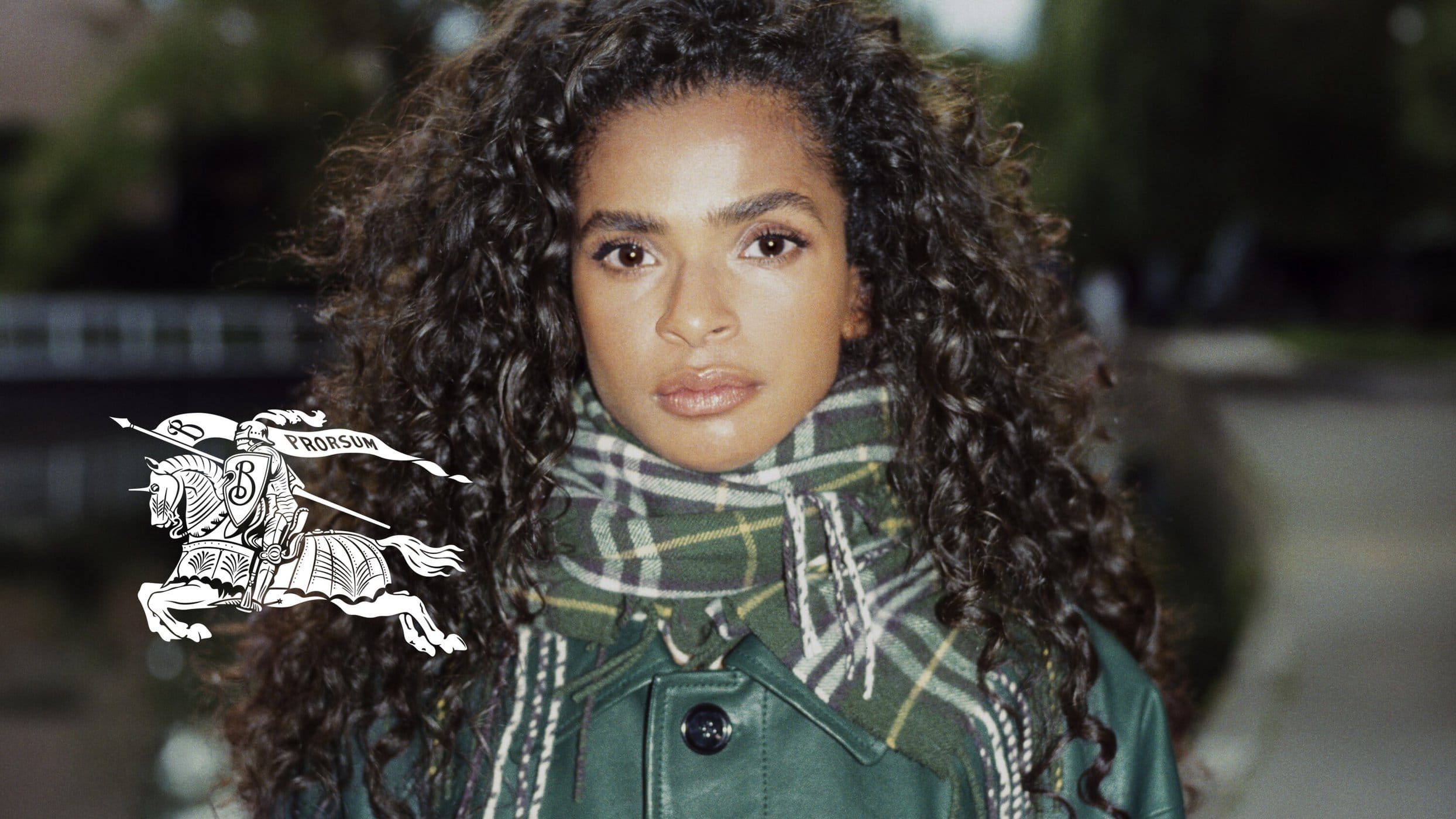 Close-up portrait of a woman with curly hair wearing a green jacket and plaid scarf. Burberry logo visible on the left side.