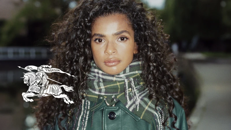 Close-up portrait of a woman with curly hair wearing a green jacket and plaid scarf. Burberry logo visible on the left side.
