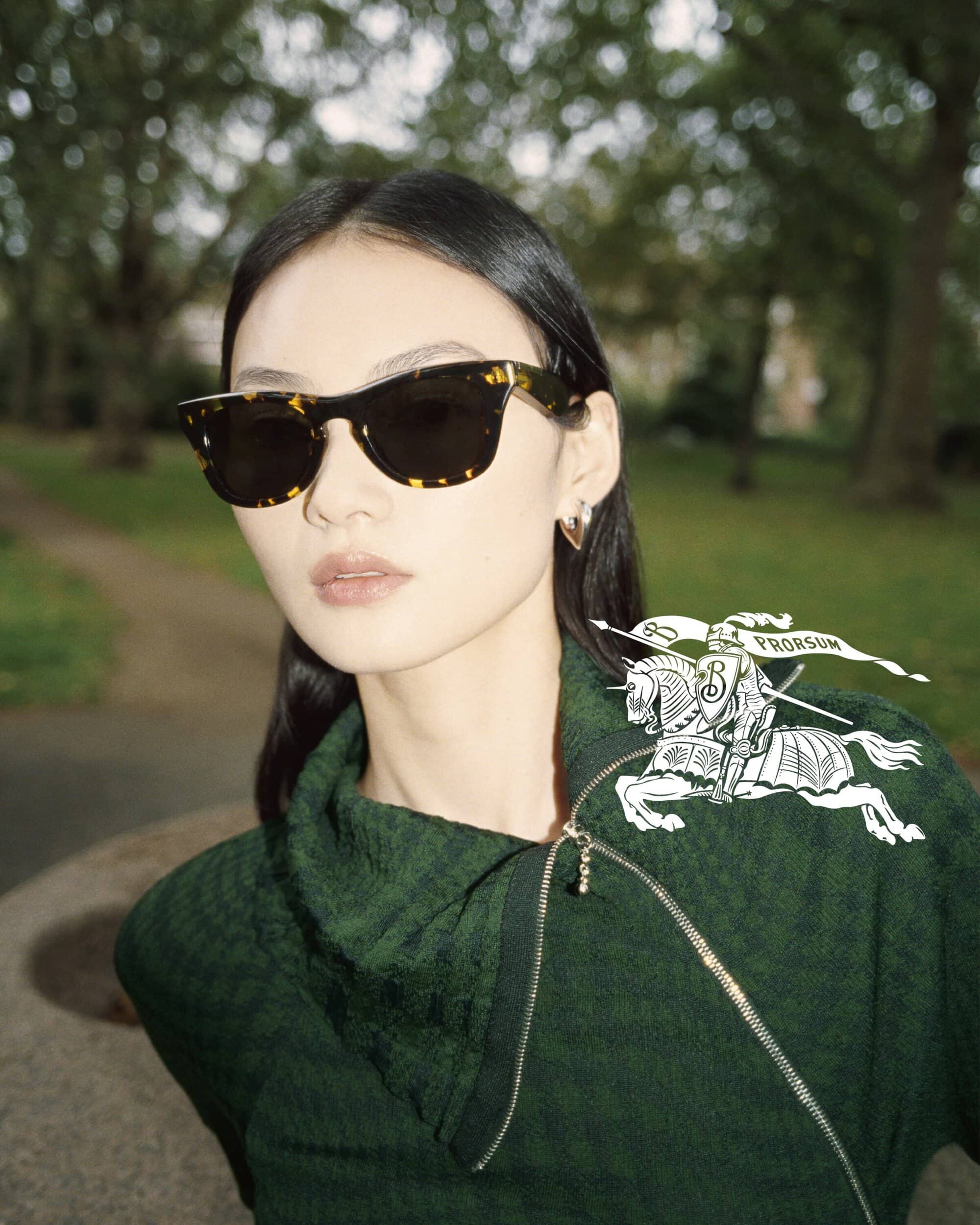 Portrait of an Asian woman wearing tortoise shell sunglasses and dark green textured top in an outdoor setting with a Burberry pin.