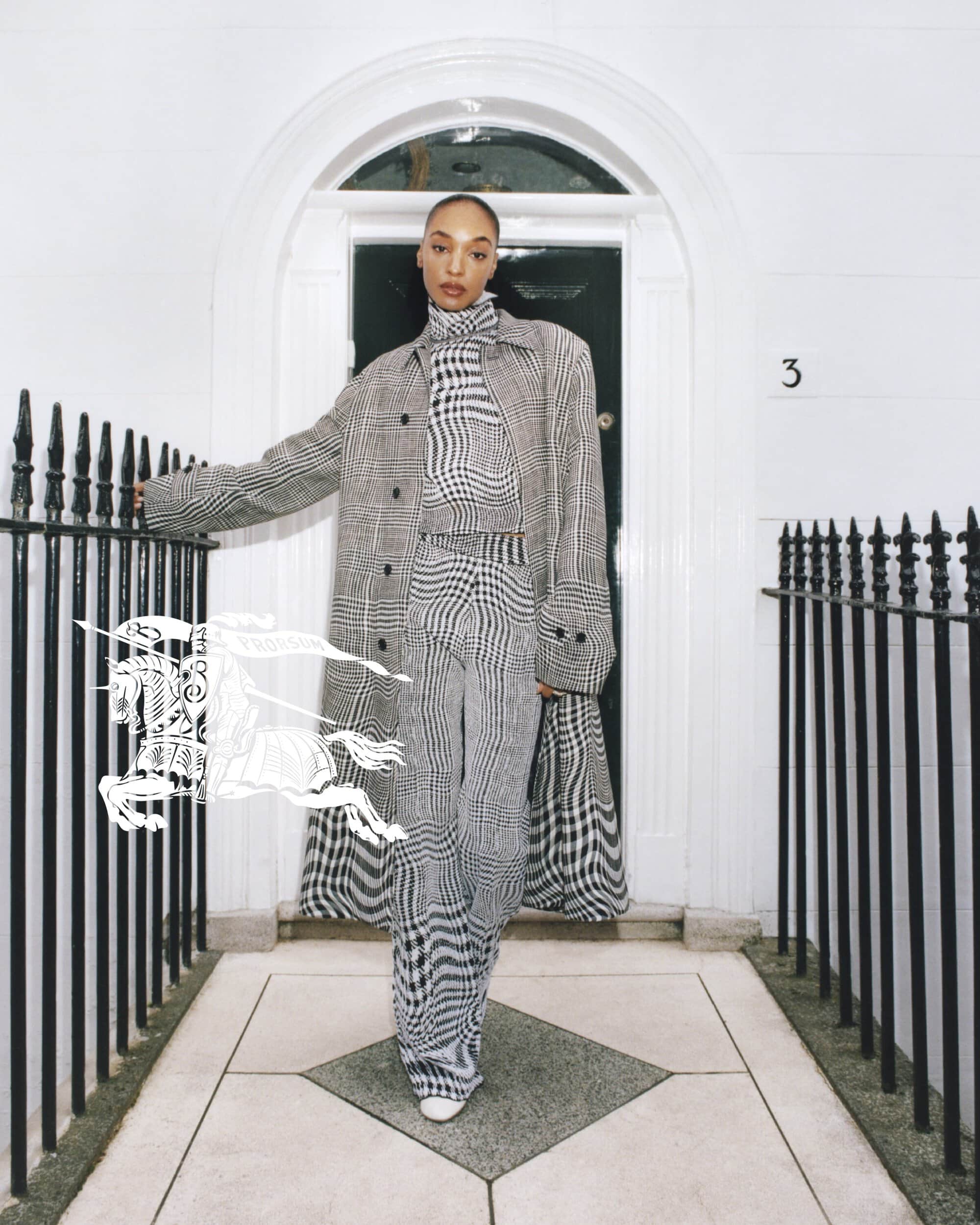 A Black female model poses in a grey plaid turtleneck, pants, and coat near a white door, showcasing a high-fashion editorial style.