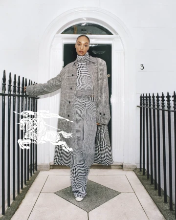 A Black female model poses in a grey plaid turtleneck, pants, and coat near a white door, showcasing a high-fashion editorial style.