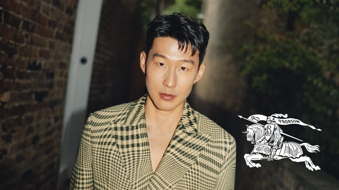 Portrait of a handsome Asian man in a checkered blazer, posing in front of a brick wall with a Burberry logo visible. Fashion shot.