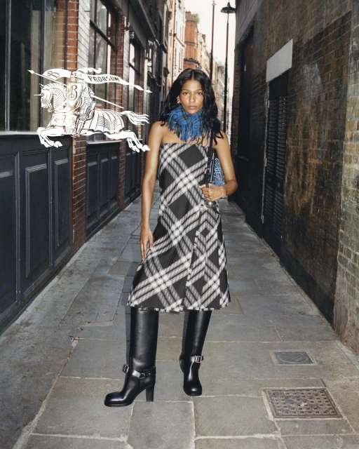 A solo model wearing a black and white plaid strapless dress with black boots poses in an outdoor alleyway with brick buildings.