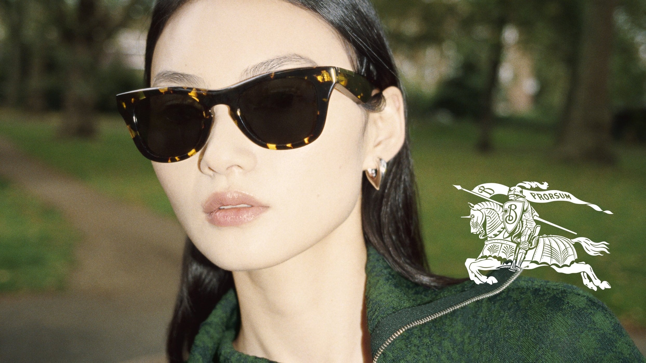Close-up of an adult woman wearing sunglasses with a green top outside. The Burberry logo is subtly placed on the garment.