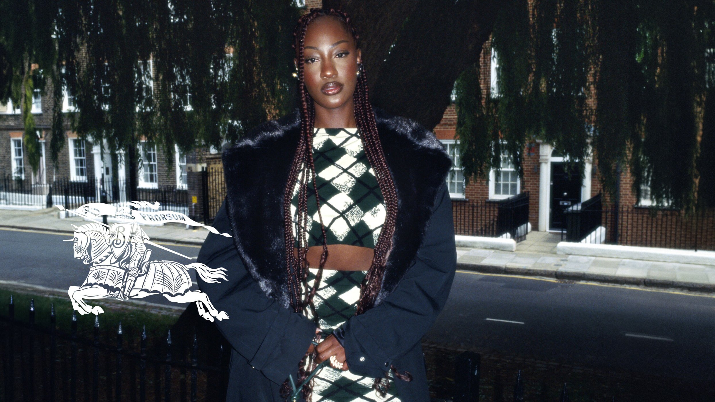 Woman in an argyle crop top and skirt posing in front of an urban backdrop, wearing a black fur-trimmed jacket, and braided hair.
