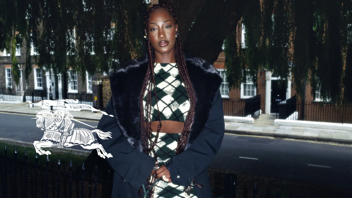Woman in an argyle crop top and skirt posing in front of an urban backdrop, wearing a black fur-trimmed jacket, and braided hair.
