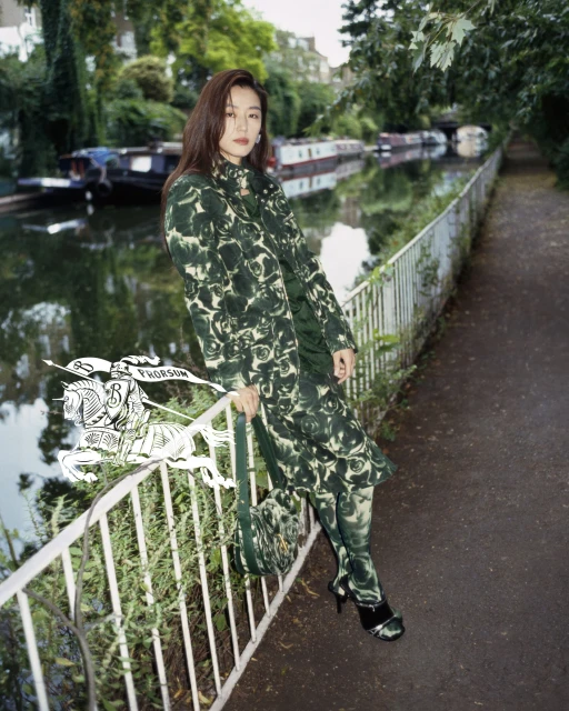 A fashionable woman poses in a printed coat, bag, and tights along a canal with boats, captured in a medium shot on a paved path.