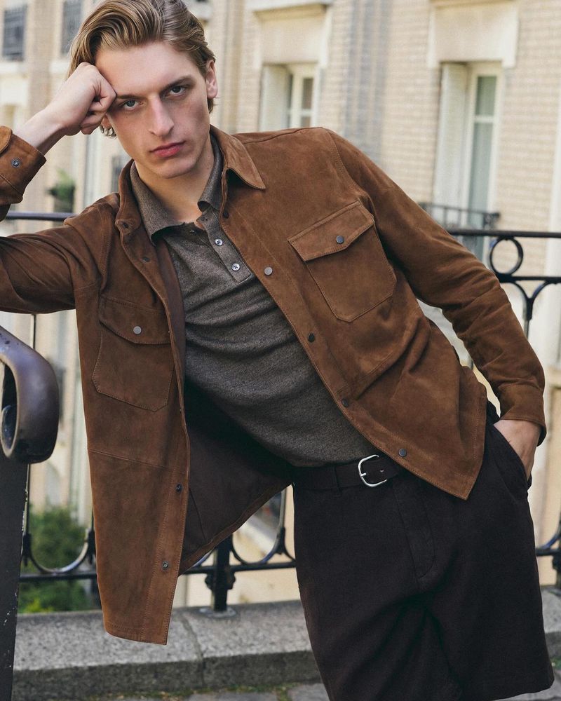 A young adult man in a brown suede shirt and grey polo shirt leans against a railing on a sunny day. Fashion editorial.