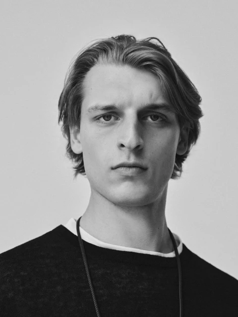 Monochrome portrait of a young adult male with medium-length hair wearing a dark sweater against a solid neutral backdrop.