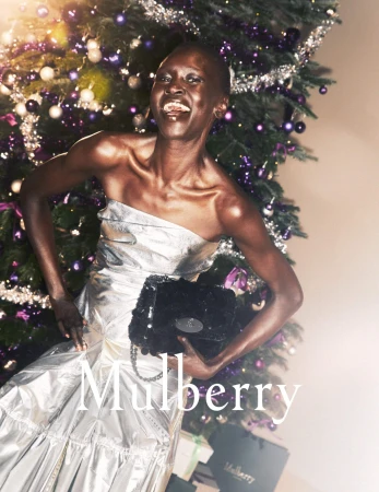 A smiling African American woman poses with a black Mulberry purse in a silver dress next to a Christmas tree with violet ornaments.