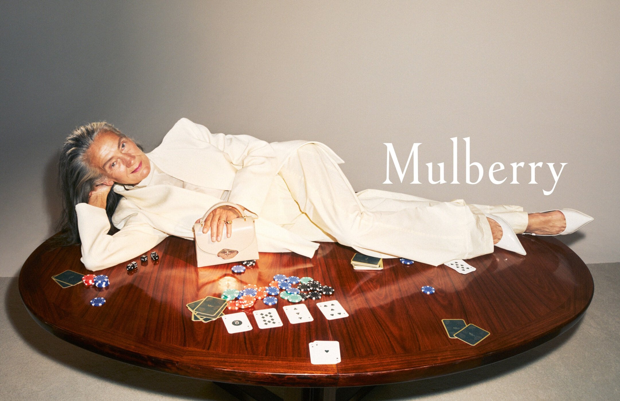 Woman in a suit poses on a poker table with playing cards, dice, and chips. The brand name Mulberry is superimposed on the image.