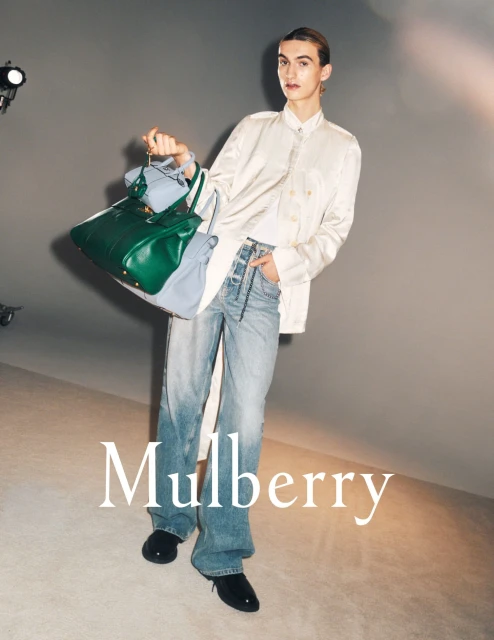 Fashion shot of a model in light-wash jeans, a white shirt and blazer, and black shoes, holding a green leather handbag.