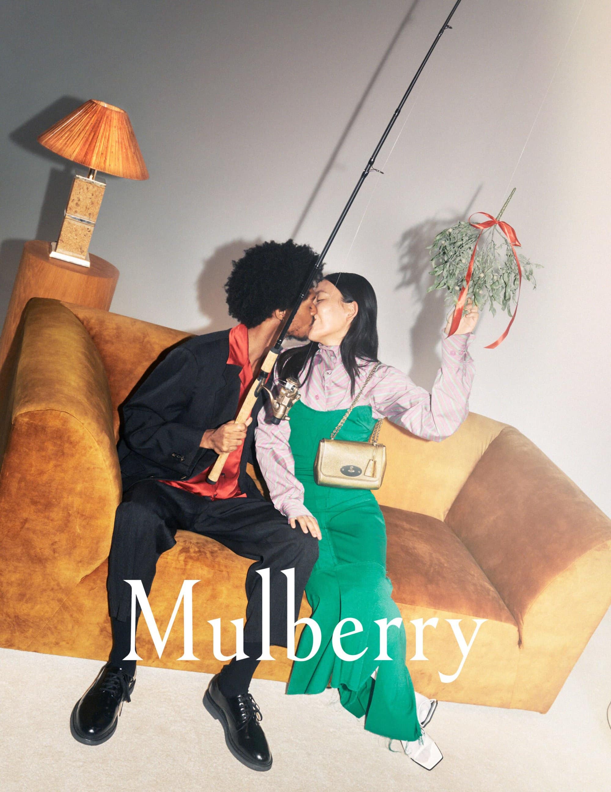 Stylish indoor studio shot features a kissing couple on a sofa, adorned with a fishing rod and mistletoe, promoting the Mulberry brand.