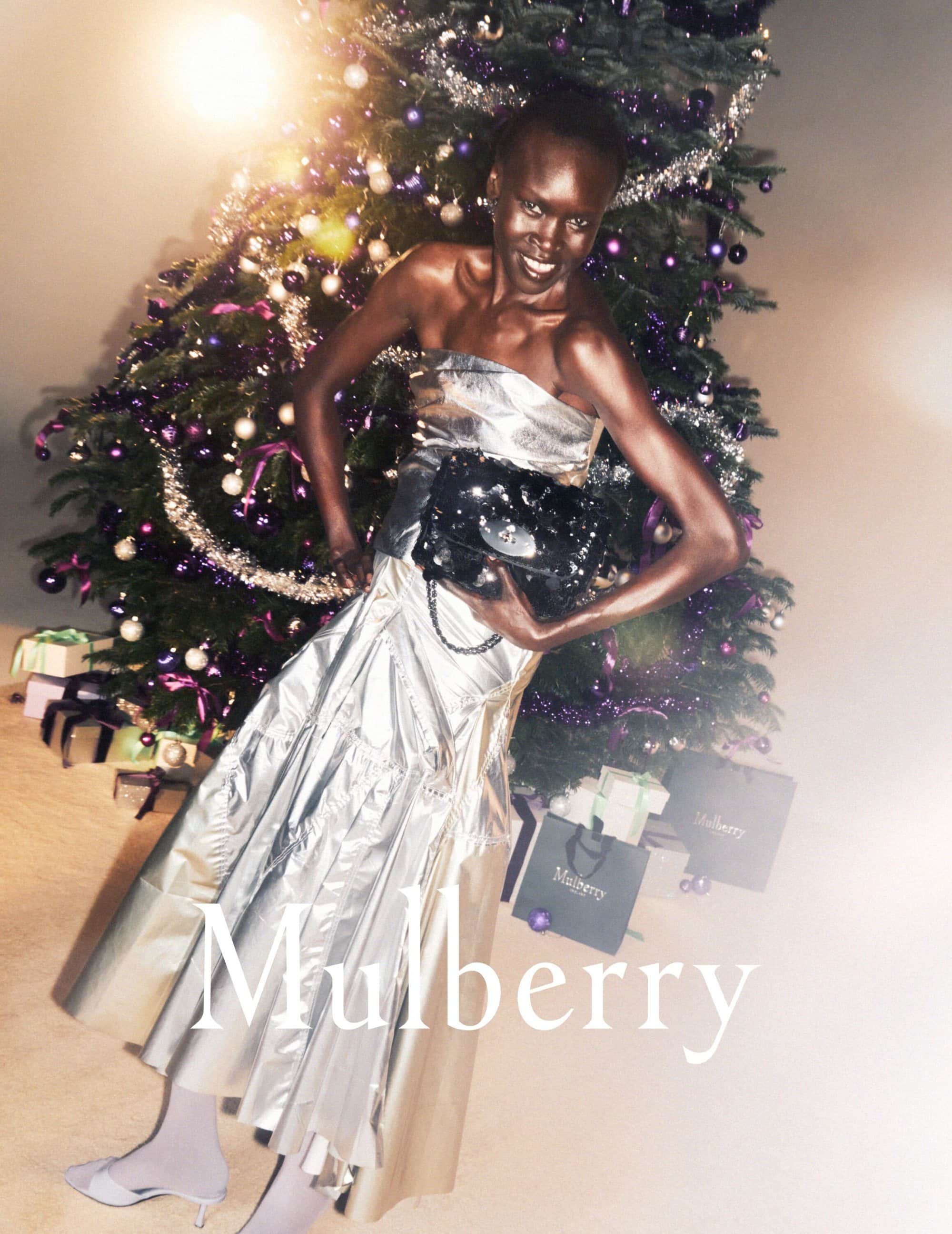 A smiling African woman poses next to a decorated Christmas tree in a silver strapless dress, holding a black patent leather bag, indoors.